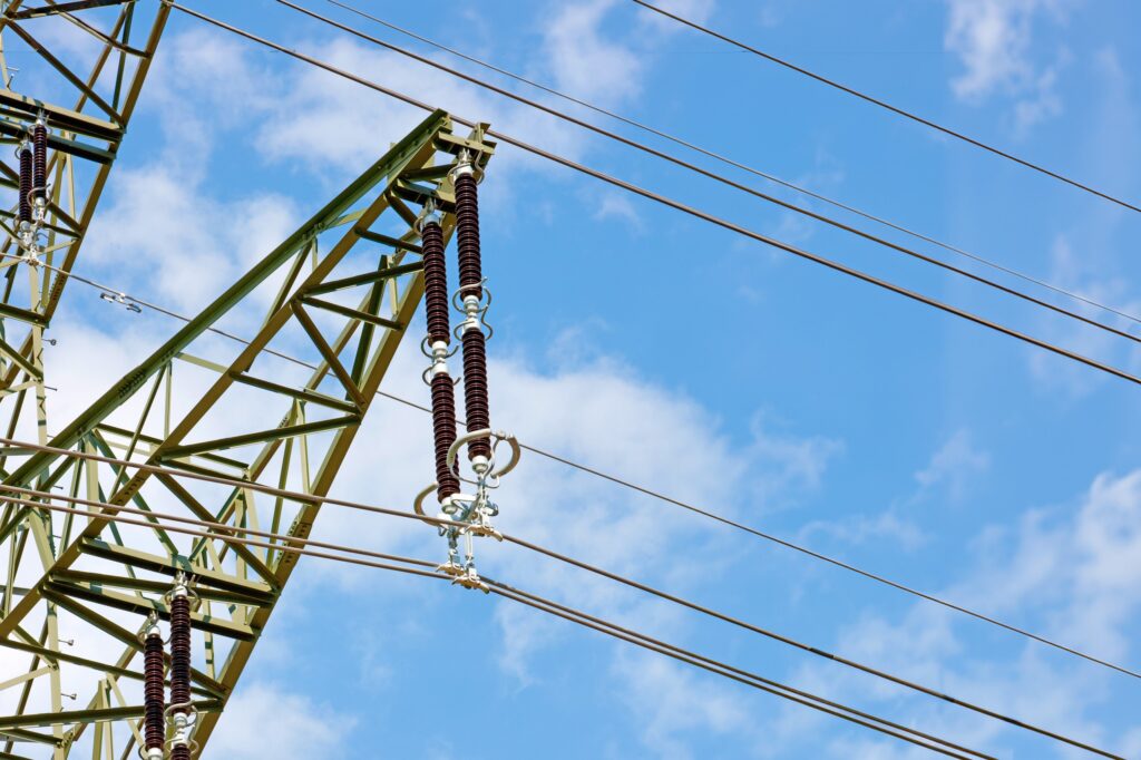 Arm eines Strommast mit Hängeketten (Isolatoren, Klemmen, Coronaringen) und Stromleitungen bei blauem, leicht bewölkten Himmel